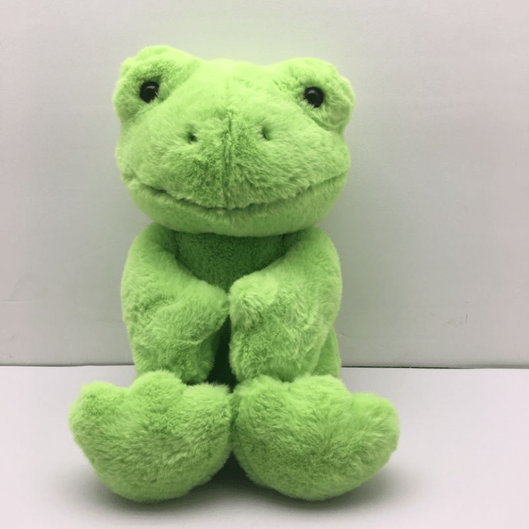 Peluche grenouille verte sur une surface blanche devant un mur blanc.